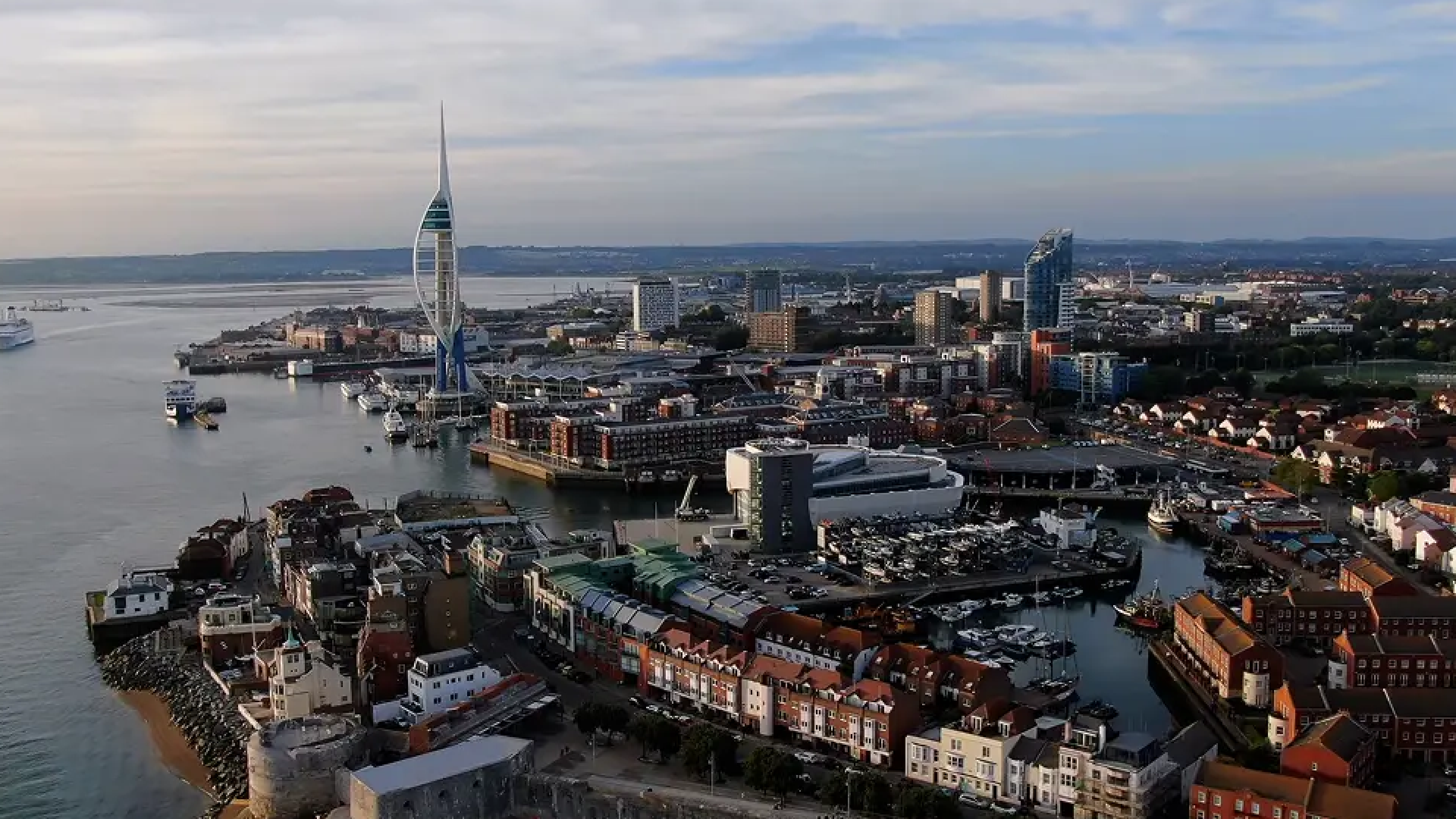 Aerial view of a coastal city featuring the Spinnaker Tower, a harbor with boats, and buildings along the waterfront.