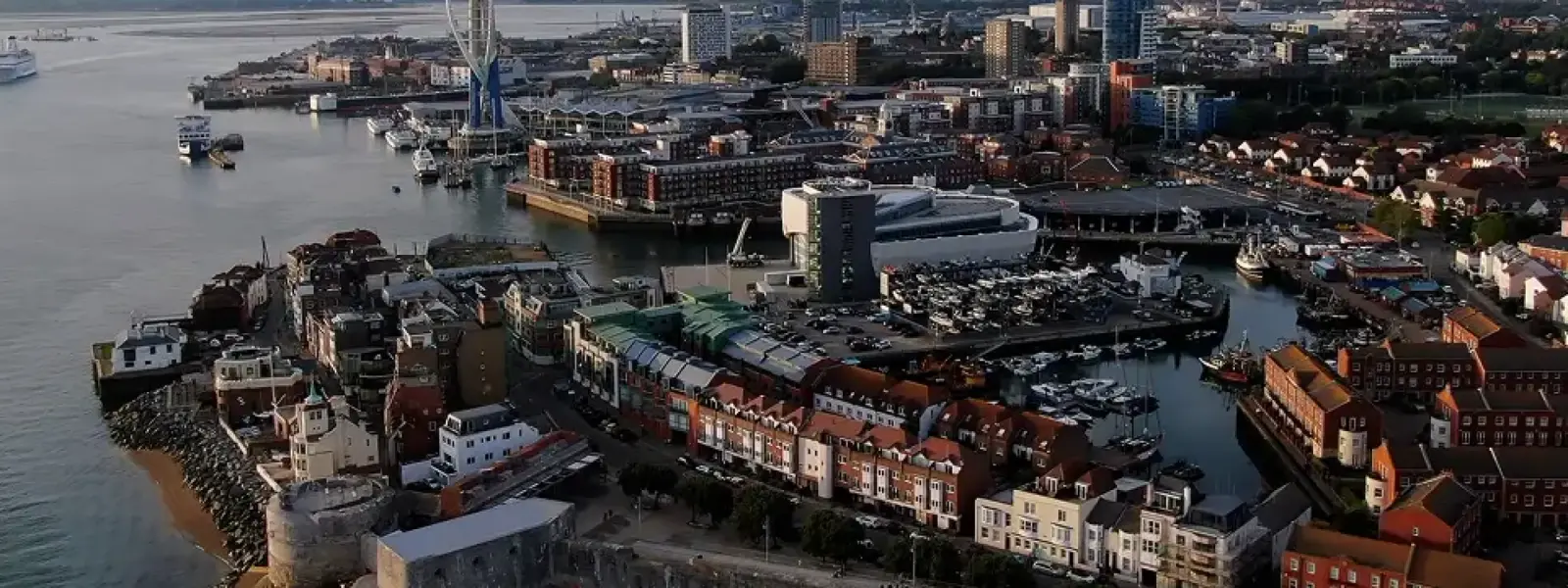 Aerial view of a coastal city featuring the Spinnaker Tower, a harbor with boats, and buildings along the waterfront.