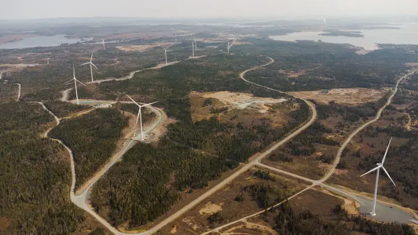 Aerial view of a wind farm with multiple turbines, winding roads, forests, and distant lakes.