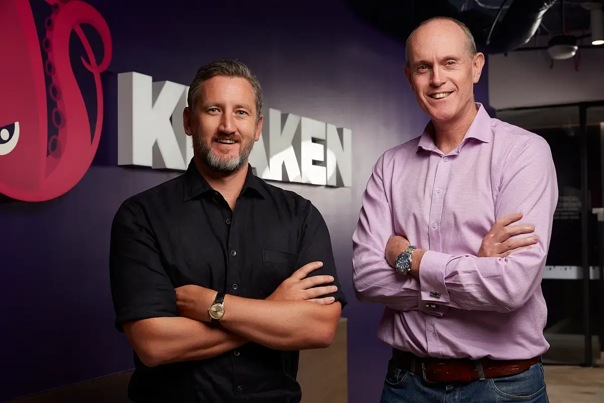 Two smiling men with crossed arms in front of a purple wall with a Kraken logo.