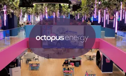 Vibrant, multi-level Octopus Energy office with pink and blue decor, hanging plants, purple lights, and people working. The Octopus Energy logo is prominent.
