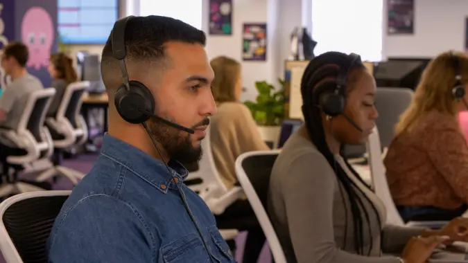 Employees wearing headsets with microphones work at computers in an office.
