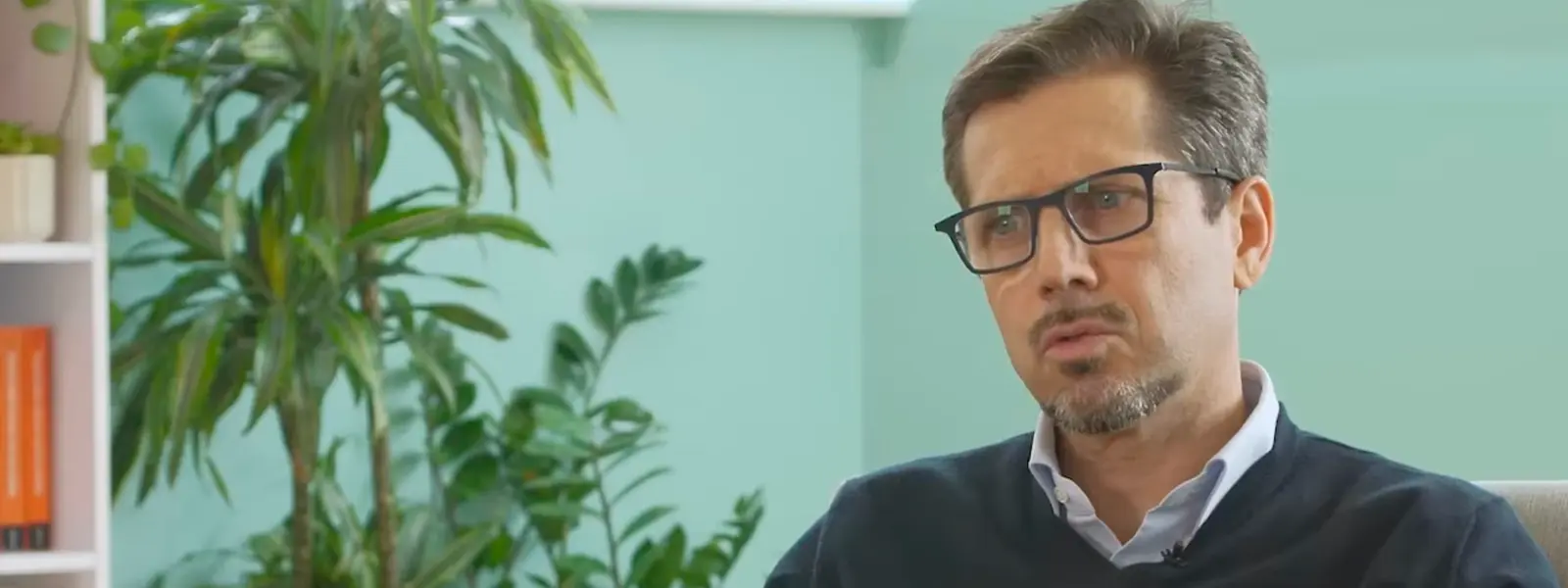 A man with glasses and a beard, wearing a navy sweater, speaks and gestures. Plants and a bookshelf are in the background.
