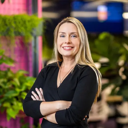 A smiling blonde woman in a black top with crossed arms stands against a blurred background of green plants.