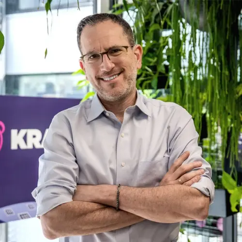 A smiling man with glasses and a light blue shirt crosses his arms in an office with plants and a purple sign.