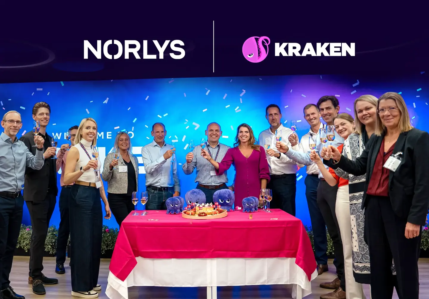 A group of people from Norlys and Kraken toast, celebrating in front of their logos with a cake and purple octopus plushies.