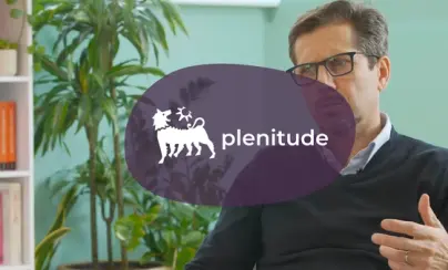 A man in glasses and a navy sweater talks, with plants, a bookshelf, and a purple Plenitude logo overlaid.