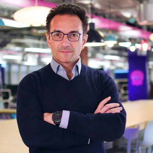 A man in glasses and a blue sweater with crossed arms, wearing a smartwatch, stands in a vibrant office.
