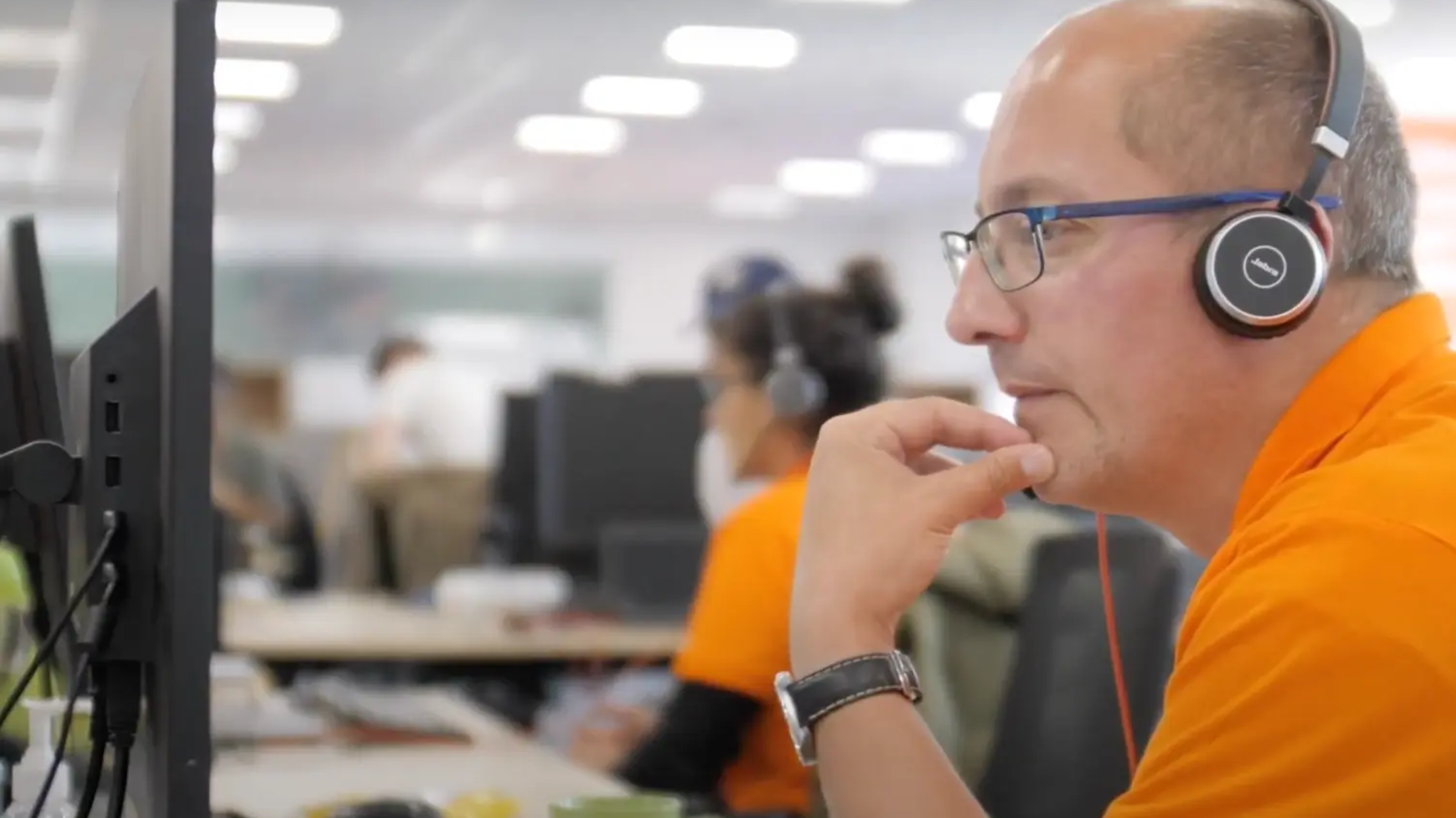 Man in orange shirt with headphones and glasses looking at a computer in an office.