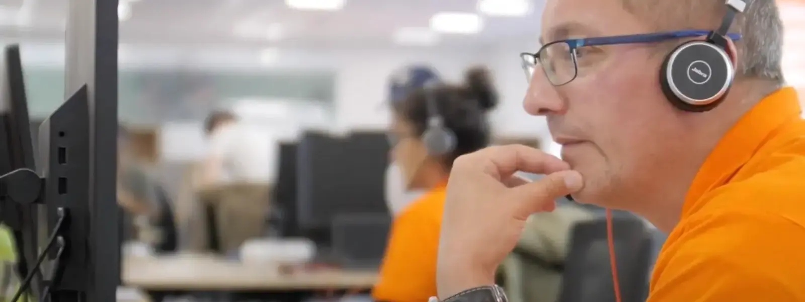 Man in orange shirt with headphones and glasses looking at a computer in an office.