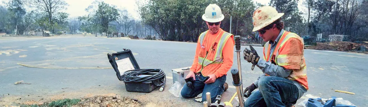 Two workers in hard hats and safety vests perform tests on a road, with fire-damaged trees in the background.