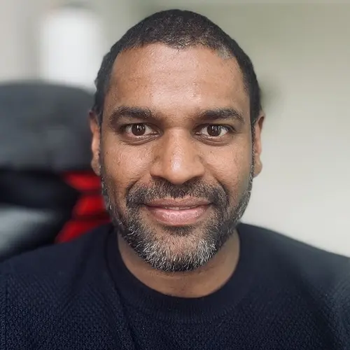 A smiling man with dark skin and a graying beard wears a dark sweater.