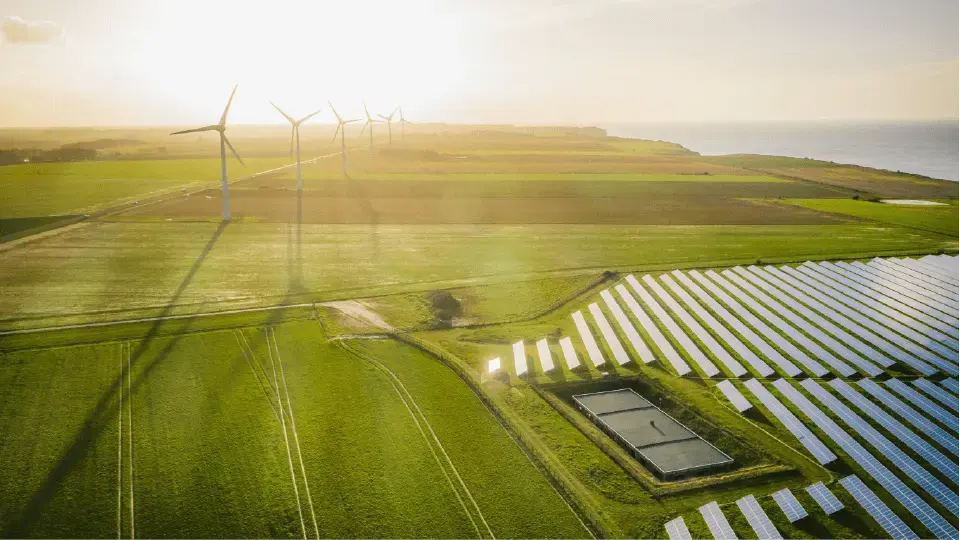 Aerial view of wind turbines and solar panels in green fields by the sea at golden hour.