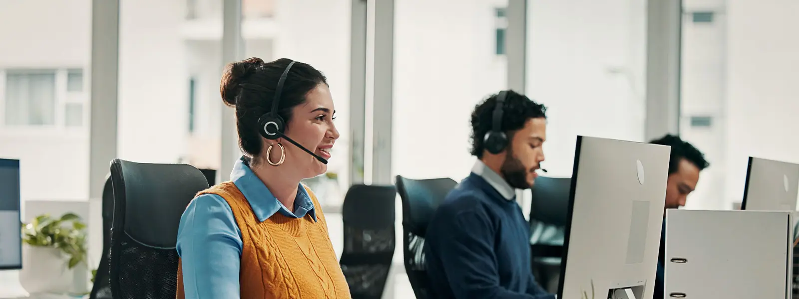 A smiling customer service agent wearing a headset and typing at a computer, with other agents working in the background.