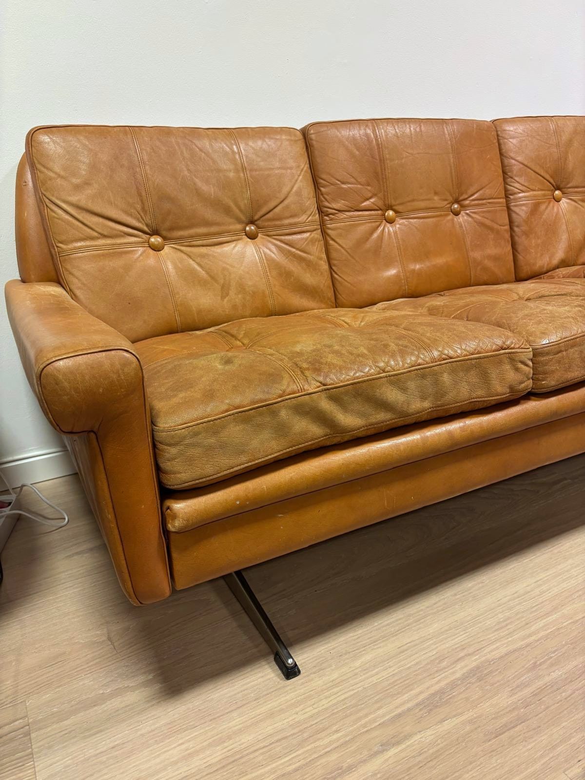 Sofaen sett skrått fra siden, med knappede ryggputer i cognacfarget skinn.