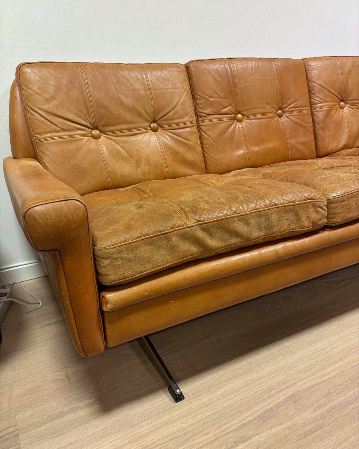Sofaen sett skrått fra siden, med knappede ryggputer i cognacfarget skinn.
