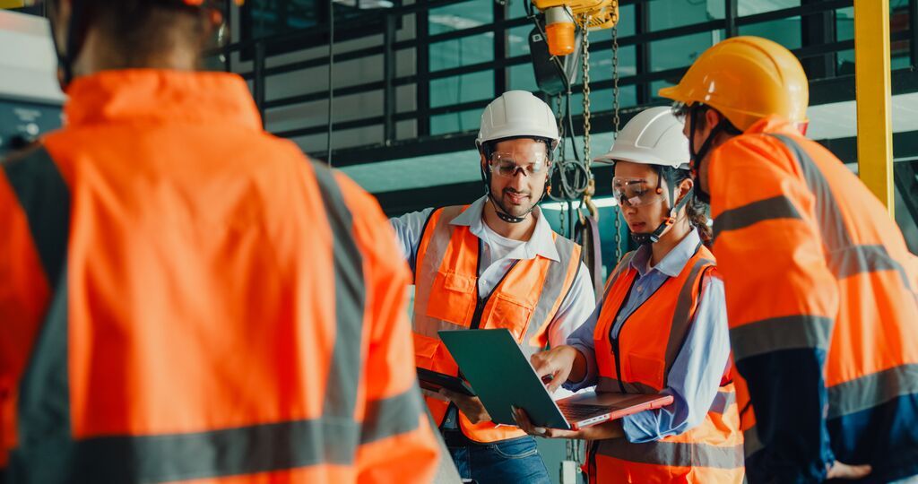 Workers in high-visibility vests and hard hats gathered around a laptop in a warehouse setting