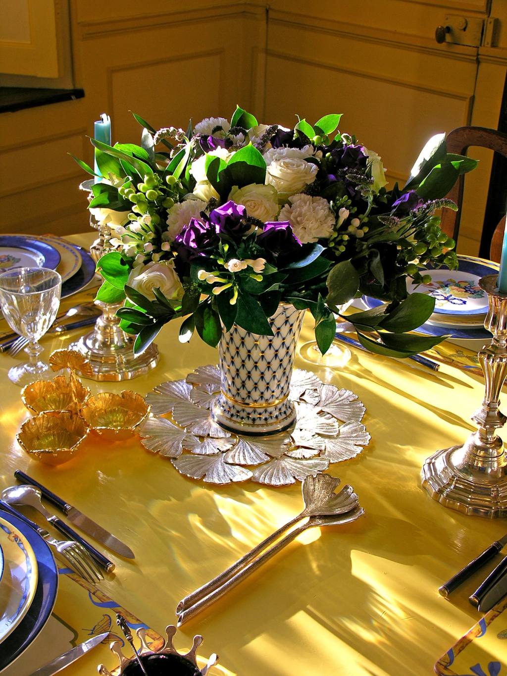 Table on a yellow cloth with white roses and purple flowers in a blue-and-white vase, silver and china