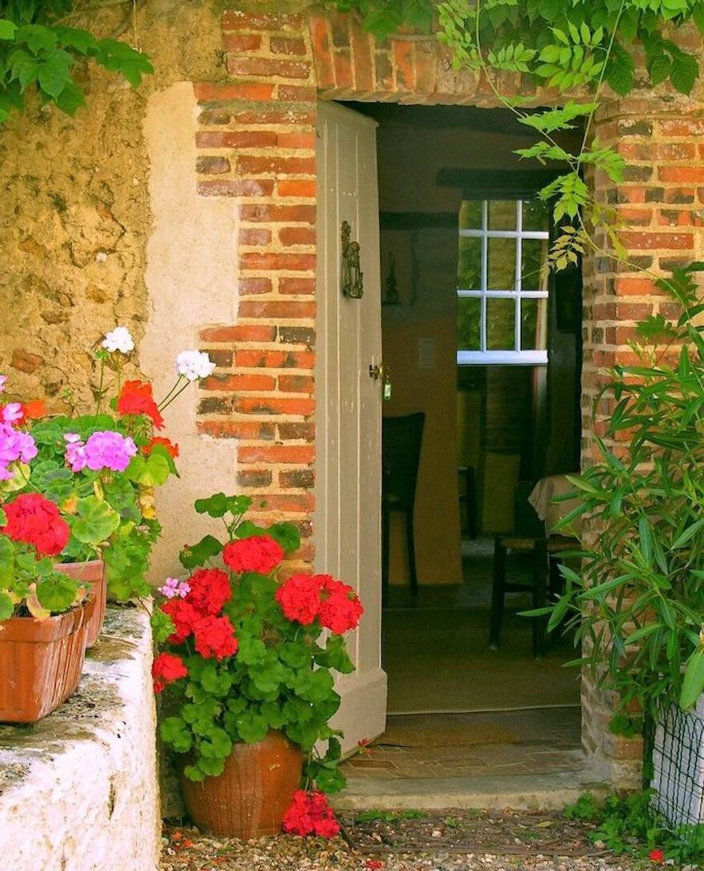 Open cottage door framed in old brick, with pots of red geraniums on the step