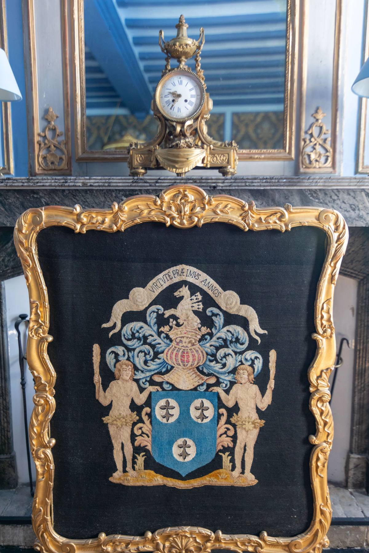 Gilt-framed fire screen embroidered with a coat of arms, clock on the mantel above