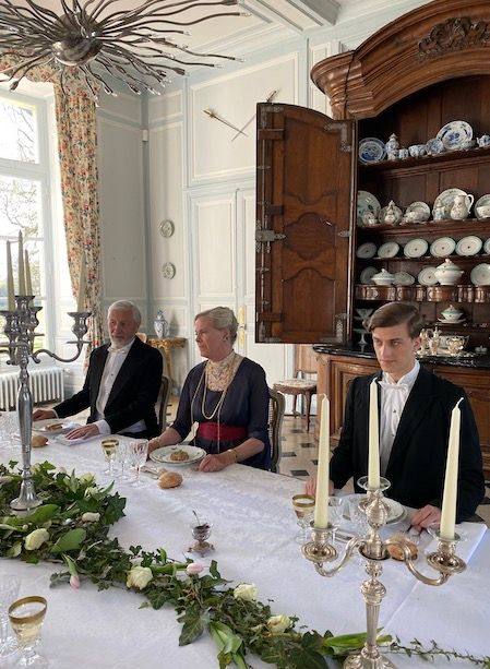 Guests in evening dress at a candlelit table with a greenery garland, before an open porcelain dresser