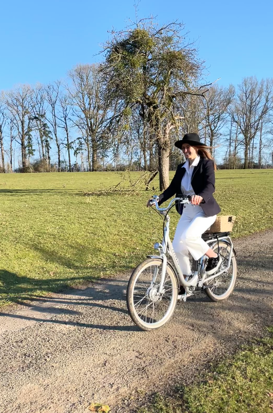 Guest in a black hat riding a white bicycle with a wicker basket along a path in the park