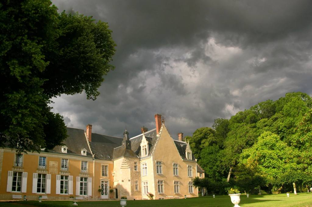 The sunlit château façade beneath dark storm clouds, lawn and trees in the foreground