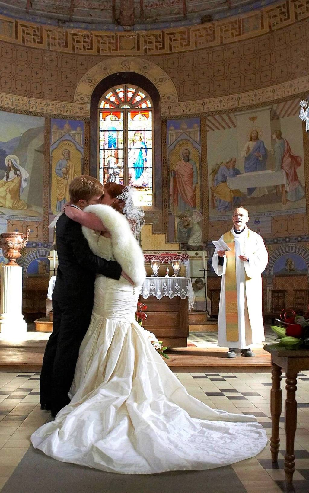 Bride and groom kissing at the altar of the church of Conflans, a priest looking on beneath painted murals