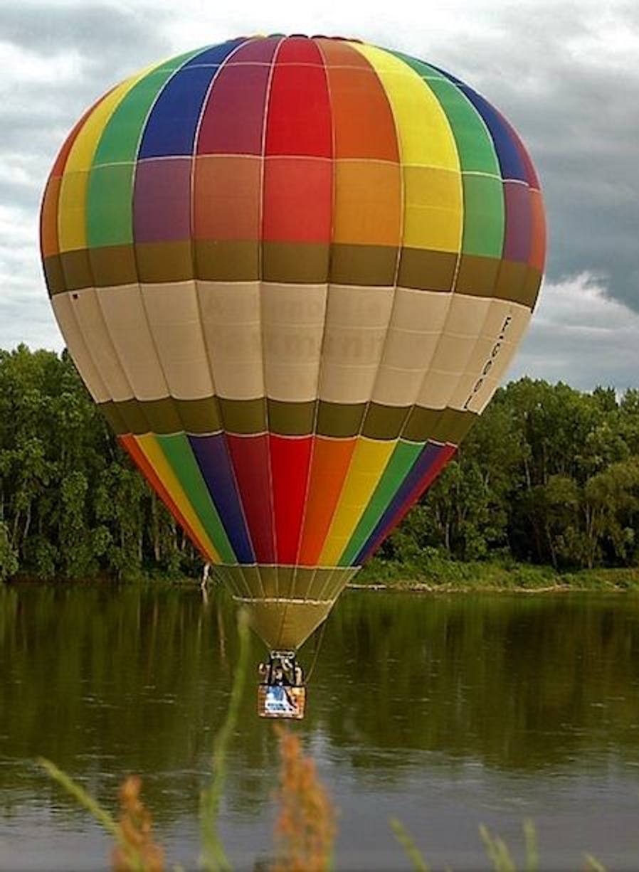 Gestreifter Heißluftballon gleitet dicht über einer von Bäumen gesäumten Wasserfläche