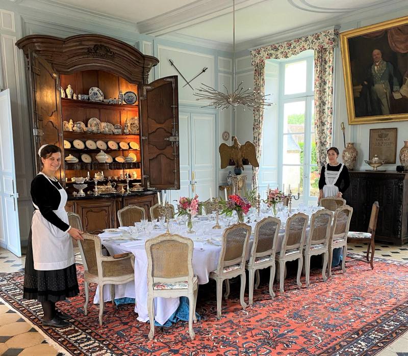 Grande Salle à Manger : longue table dressée, vaisselier et deux serveuses en robe noire et tablier blanc