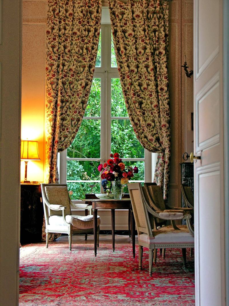 Le Grand Salon par une porte ouverte : rideaux fleuris, haute fenêtre, roses sur une table, tapis rouge