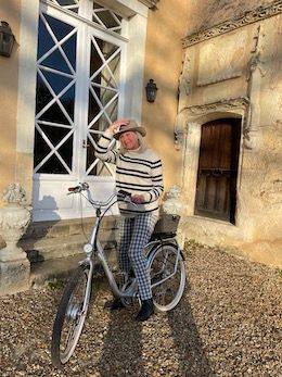 Une femme en pull rayé et chapeau pose avec un vélo devant la porte vitrée du château