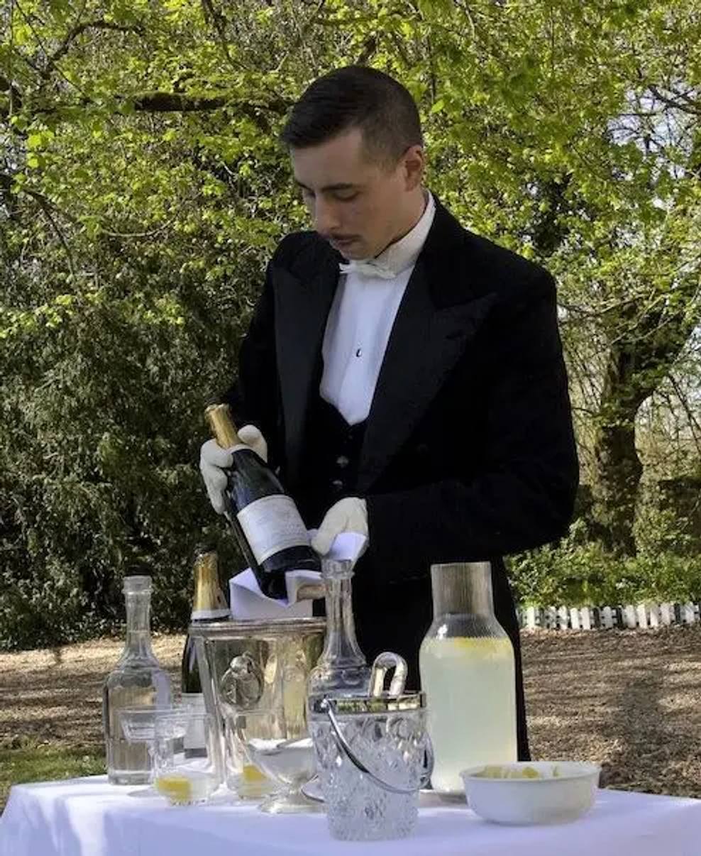 Butler in tails and white gloves pouring wine at a garden-party table under the trees