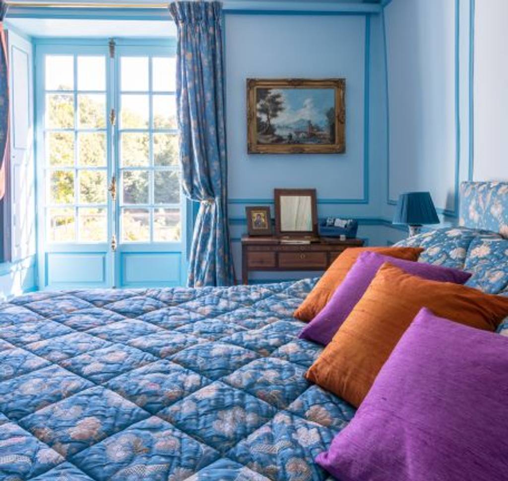 Chambre Turquoise: blue quilted bed with orange and purple cushions, French window onto the trees