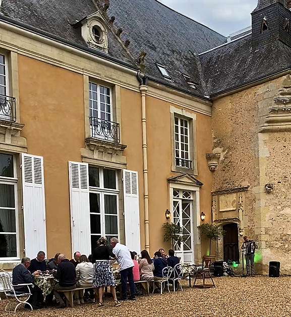 Guests dining at long tables in the courtyard before the château façade, a musician playing nearby