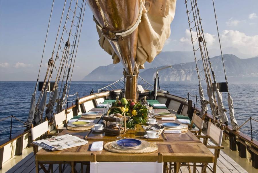 Table dressée sur le pont d'un voilier, avec la mer et les montagnes au loin