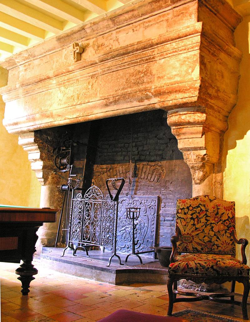 Imposante cheminée de pierre aux consoles sculptées, croix sur le manteau et fauteuil en tapisserie