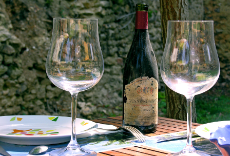 Eine alte Flasche Vosne-Romanée zwischen zwei Weingläsern auf einem Tisch im Freien vor einer Steinmauer