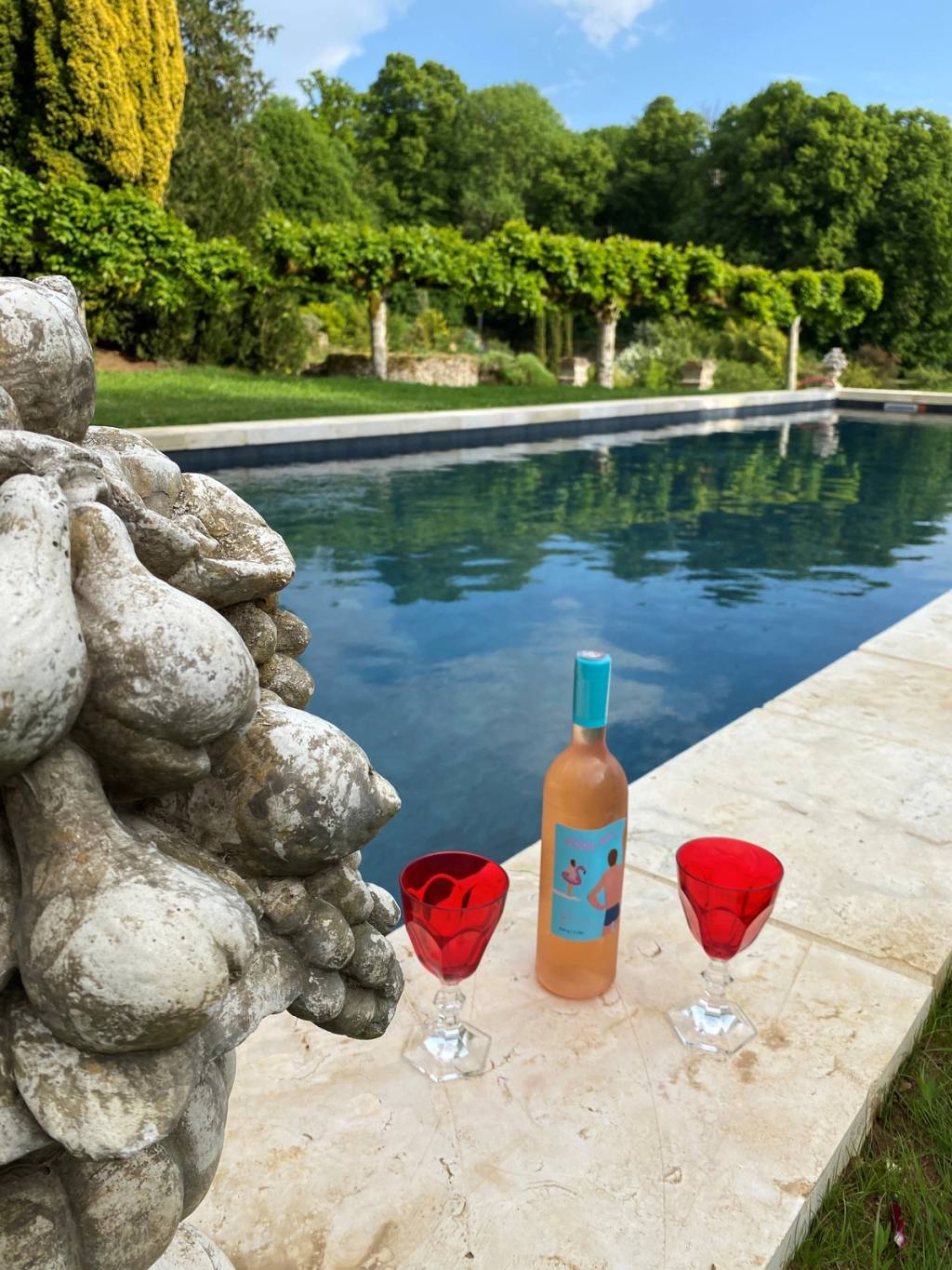Flasche Rosé und zwei rote Gläser am Poolrand, dahinter der Garten
