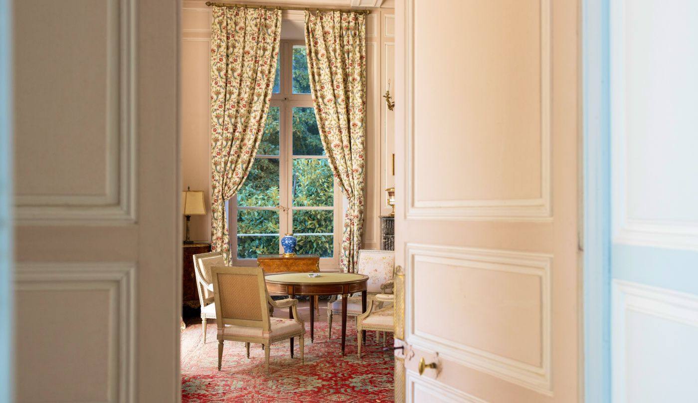 Salon vu par une porte à double battant : table ronde, chaises et rideaux fleuris