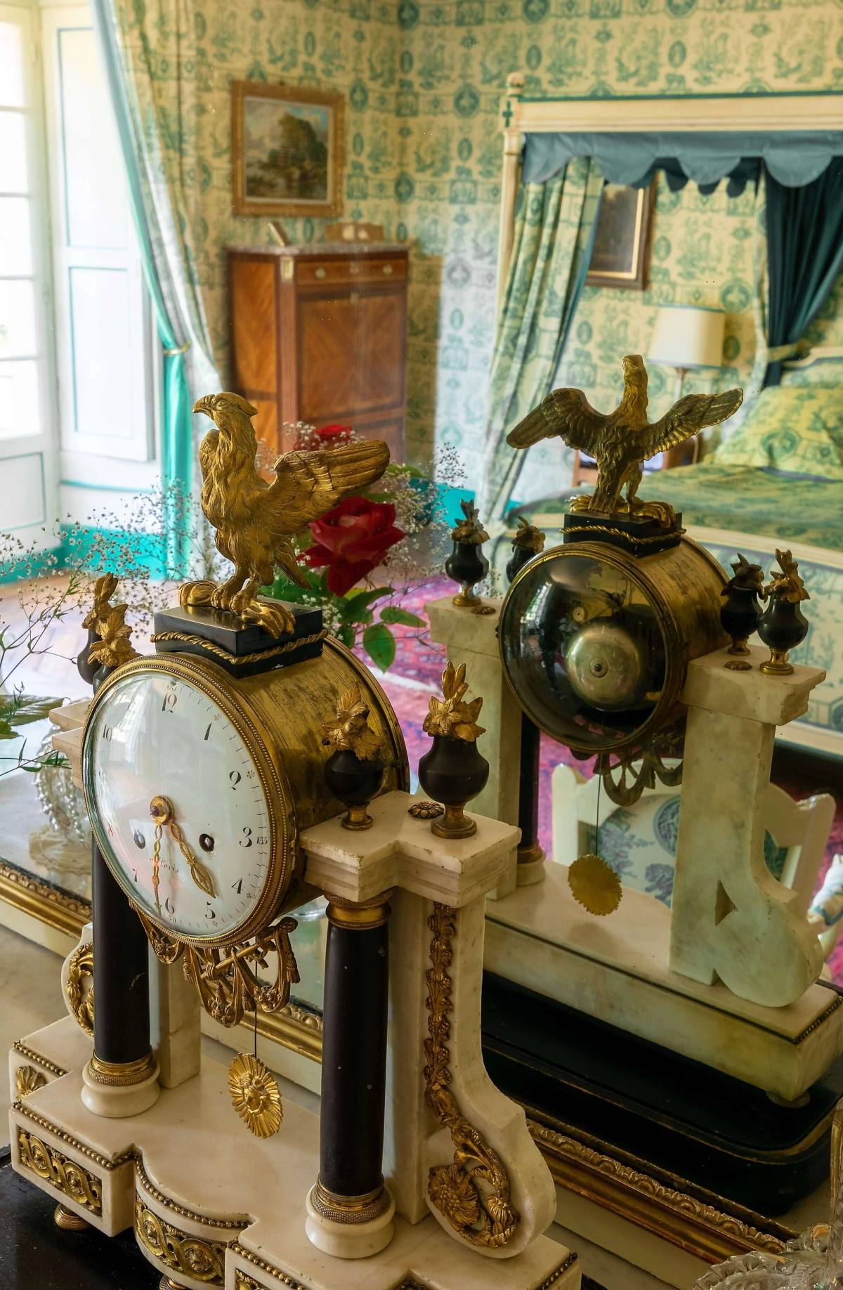Marble and gilt bronze mantel clocks crowned with eagles, bed reflected beyond, Chambre Esprit de Jouy Verte
