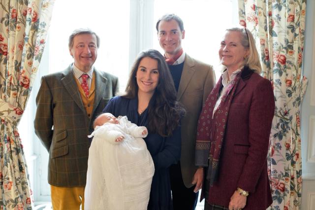 Family portrait of two couples and a baby in a long white gown, before floral curtains