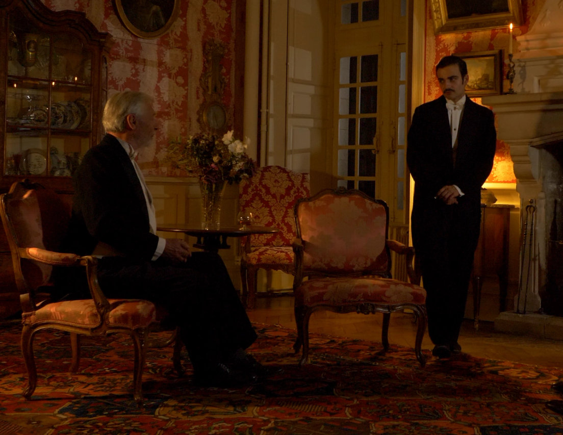Salon éclairé à la lampe, un homme assis dans un fauteuil et un autre debout en costume sombre