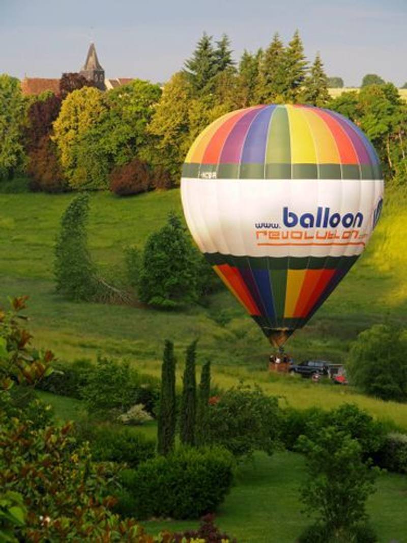 Montgolfière multicolore flottant au ras d'un vallon verdoyant, un clocher parmi les arbres