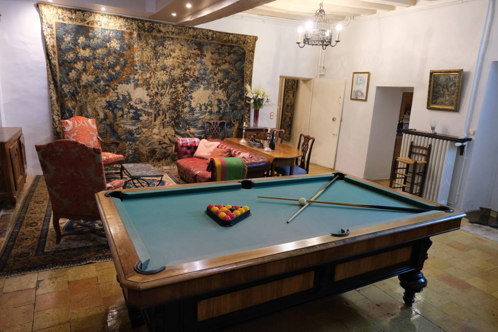 Salle de billard avec grande tapisserie de verdure, canapés rouges et lustre, sol en tomettes