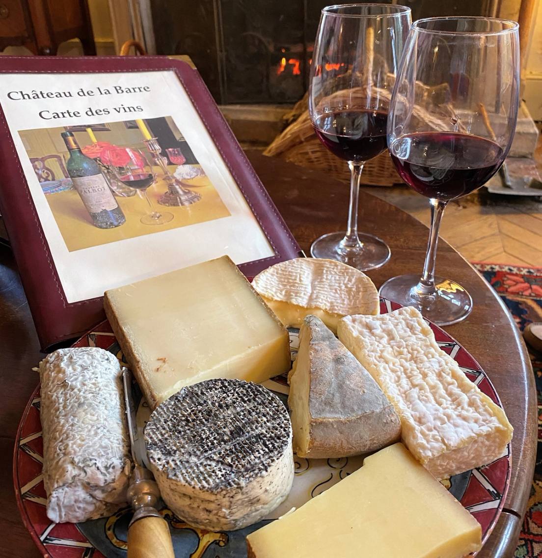 Plateau de fromages, deux verres de vin rouge et carte des vins du Château de la Barre, feu en fond