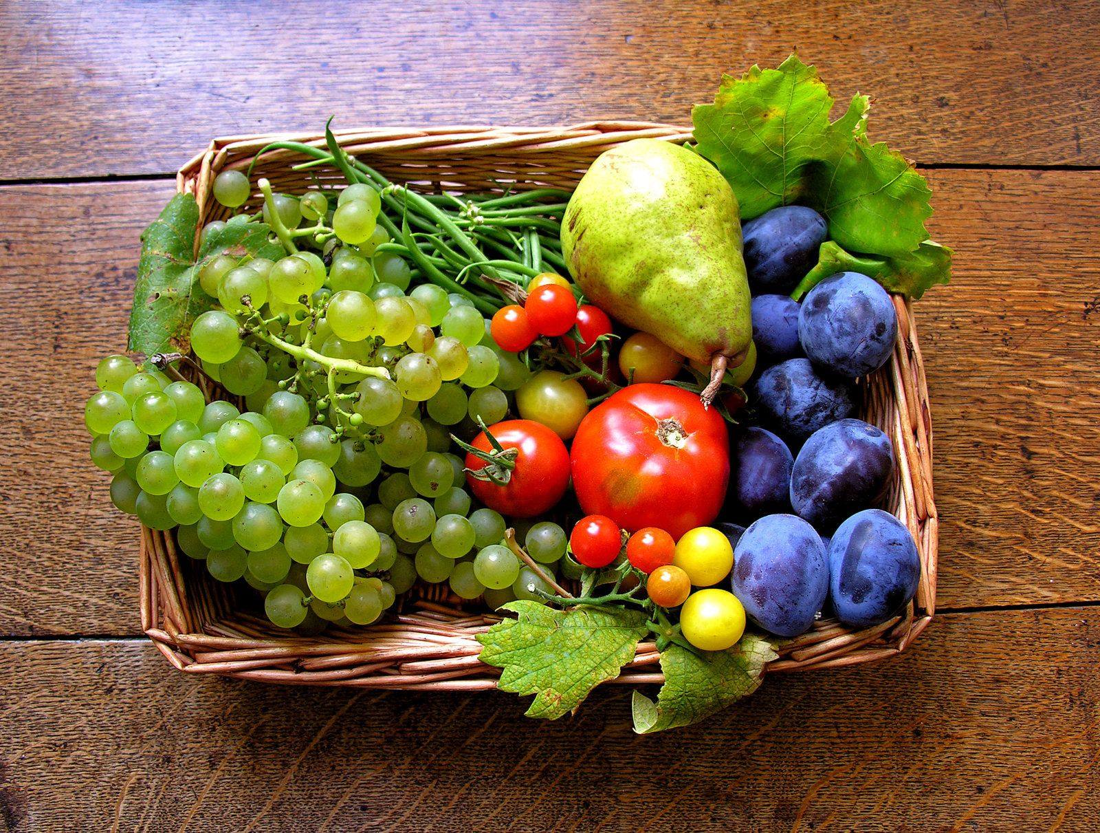 Wicker basket of organic produce: white grapes, tomatoes, a pear and plums