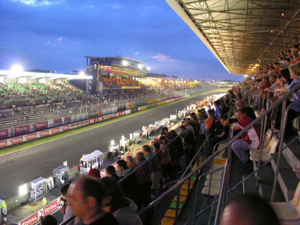 Spectateurs dans une tribune couverte surplombant un circuit automobile et ses stands au crépuscule