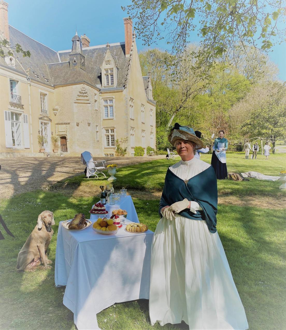 Une femme en costume du XIXe siècle près d'une table de goûter au jardin, un chien à côté, le château en fond