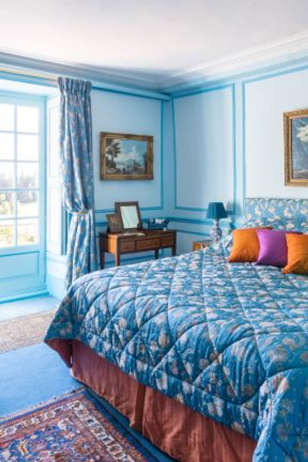 Chambre Turquoise: blue quilted bed with bright cushions, landscape paintings and a French window